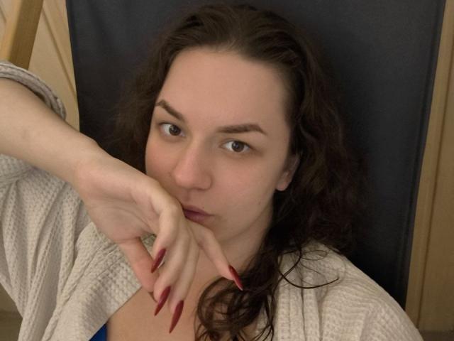 Dreia with curly brown hair, pale skin, and red nails, biting her index finger, wearing a white waffle robe, showing her small tits and subtle pout.