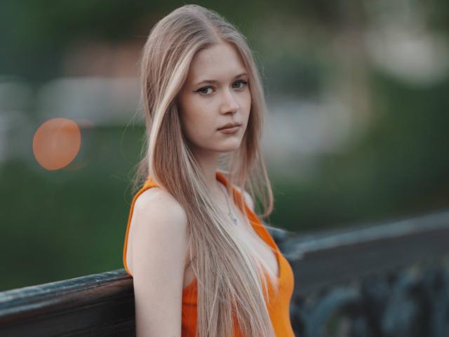 LillianaWhang has long, straight blonde hair, wearing an orange tank top, with her tits slightly visible, leaning against a black iron railing, looking serious and attractive.