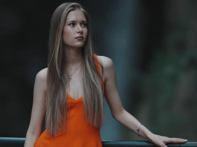 LillianaWhang with long blonde hair, orange dress, small tits, slim, looking up, hand on railing, silver necklace, bracelet, soft light, serious expression, blurry dark background.