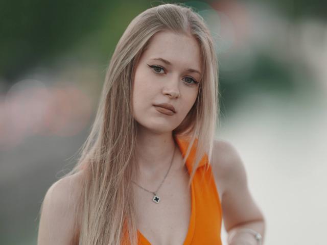 LillianaWhang has long blonde hair, green eyes, and is wearing an orange dress with a deep V. She's looking slightly to the side with a neutral expression.