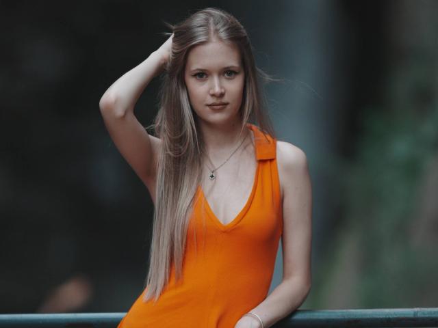 LillianaWhang with long blonde hair, wearing an orange dress, hand in hair, looking serious, small tits, slim, necklace, sitting on a green railing, blurred background.