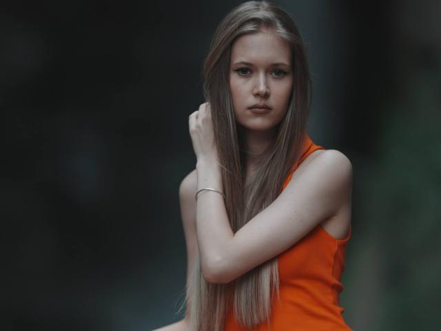 LillianaWhang with long blonde hair, wearing an orange sleeveless top, has a serious expression, her hand touching her hair, with a slim bracelet on her wrist.