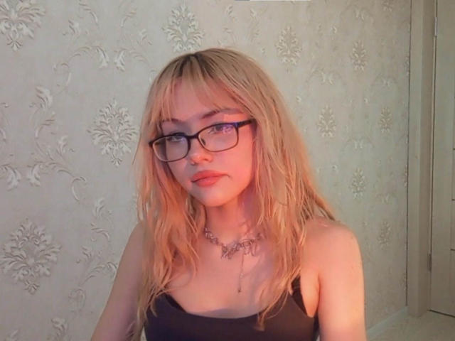Eilene Szala, com cabelo loiro, óculos e uma blusa preta, que mostra seus pequenos seios, e usando um colar, tem um tom de pele claro e uma expressão fofa e séria..