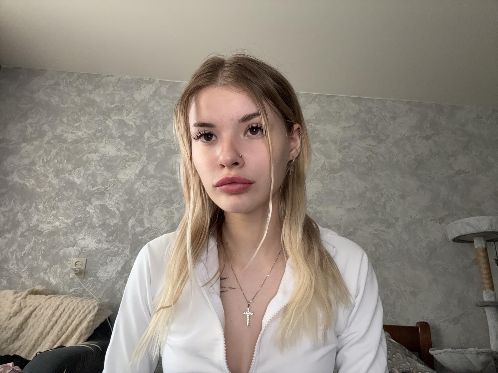 Myrtle Lilien, rubia, con una chaqueta blanca con cremallera, pechos pequeños, un piercing en la nariz, auriculares, una ligera mueca y un collar con una cruz, mirando a la cámara en una habitación gris con textura..