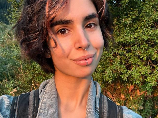 TameikaMacclellan has short brown wavy hair, brown eyes, a nose ring, and a lip piercing with a small ball. She's wearing a denim shirt and black backpack.