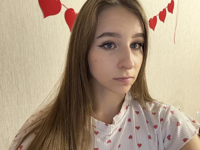 DainePoskey has long brown hair, green eyes, and a serious expression. She's wearing a white shirt with red heart patterns. Red heart decorations are above her head.