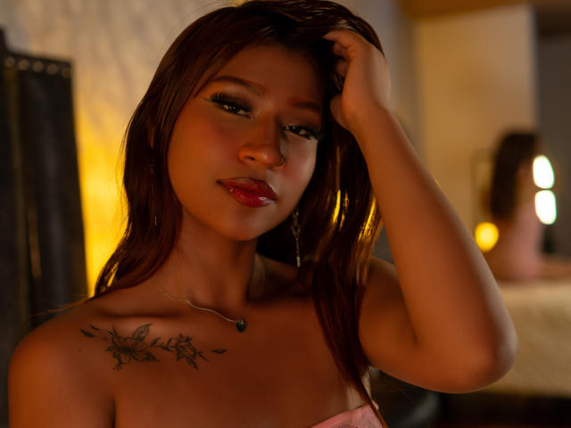 BonnieBodn with wet brown hair, red lipstick, and a floral tattoo on her right shoulder, touching her head. She's wearing a pink top, looking seductively at the camera.