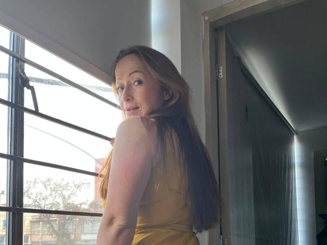 GianineLarose has long brown hair, yellow dress, and a cute smile, looking over her shoulder with sunlight on her fair skin and a door in the background.