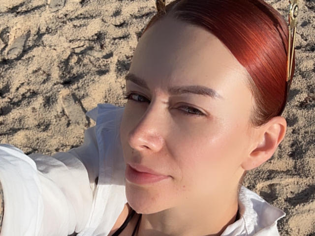 Leahred has red hair, a shiny face, and a slight smile, wearing a white shirt, lying on sandy ground with a black necklace, and sunlit skin.