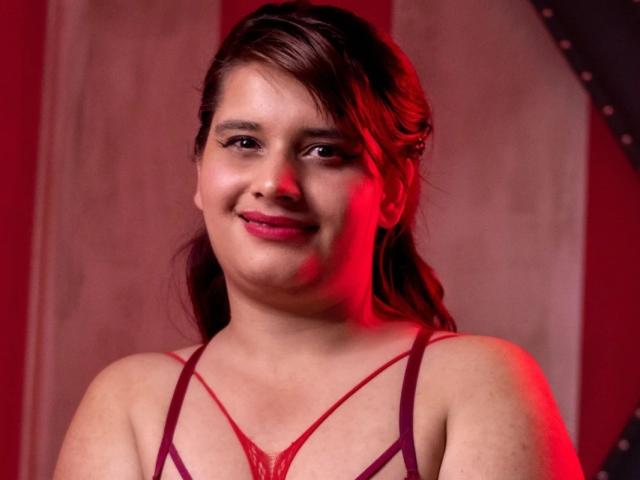 VivianFranco with brown hair, red lipstick, and a slight smile, wearing a red lace bra with thin straps, looking at the camera, red lighting on her right side.