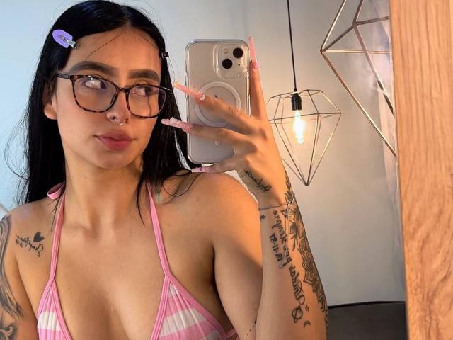AndyTaj with glasses, pink striped bikini top, tattoos, nose ring, holding phone, mirror selfie, long black hair, pink hairclip, light skin, soft smile, visible breasts, nail art.