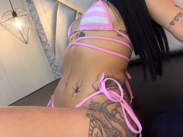 AndyTaj's pink and white striped bikini with pink straps, showing a side view of her tits, belly, and tattoos, including a spider and floral designs on her lower belly.