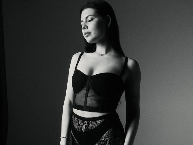 JoeyBickart with closed eyes, wearing a black lace bra and mesh skirt, has a necklace, long dark hair, and a bracelet, standing in a dark, moody black-and-white photo.