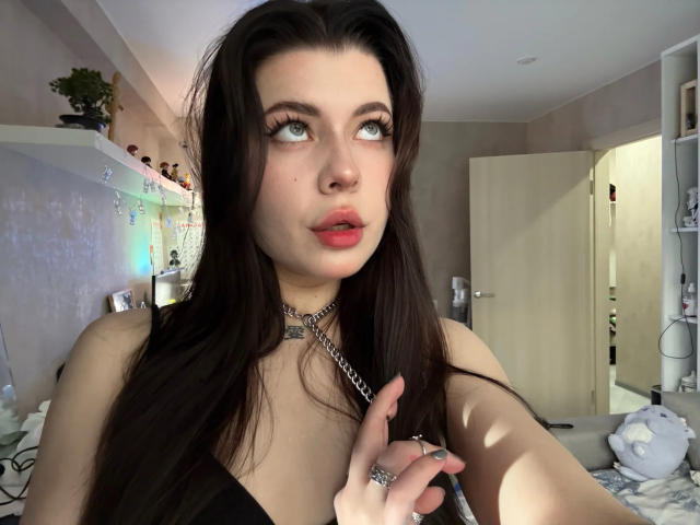 JoeyBickart heeft lang, donkerbruin haar, groene ogen en rode lippenstift. Ze draagt een zwarte top met een ketting en kijkt omhoog met een lichte frons. Borsten zichtbaar..
