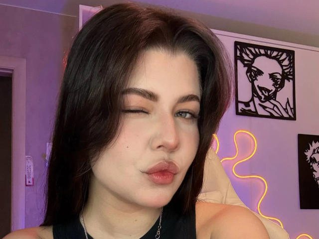 JoeyBickart has dark brown hair, pale skin, and is winking with puckered lips, wearing a black top, with a neon outline in the background and black wall art.