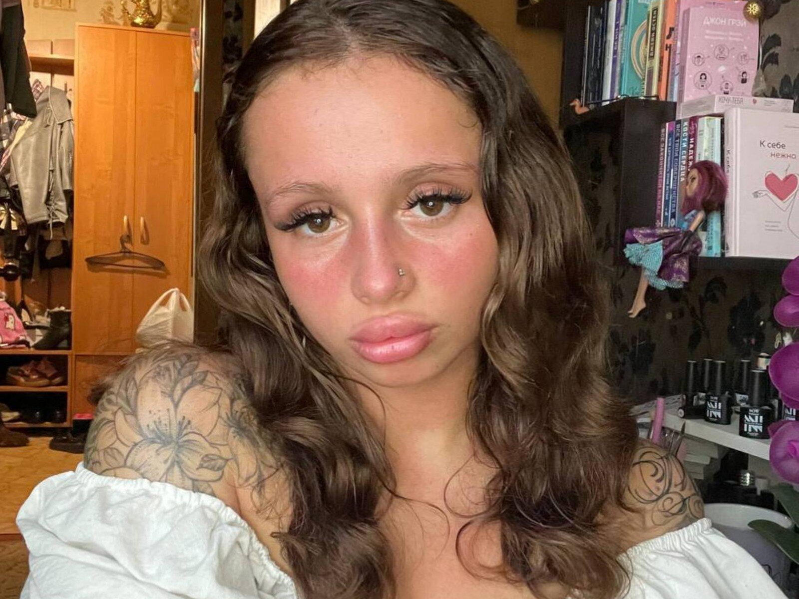 MalisaKarow, com cabelo castanho ondulado, piercing no nariz, ombros tatuados, lábios rosados e delineador, vestindo uma blusa branca com um ombro descoberto, em um quarto com prateleiras de madeira e escuras..