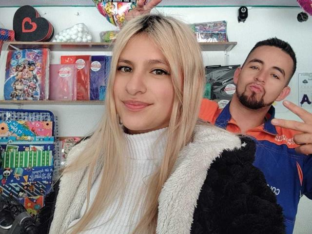 LilaHt with blonde hair, white turtleneck, and black fluffy jacket, smiling, in front of a guy with short dark hair, orange shirt, making a pouty face, holding up a peace sign.
