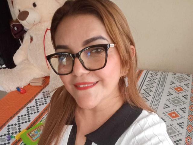 Ladynor Monroy hat braunes Haar, eine Brille mit schwarzem Rahmen, roten Lippenstift und ein süßes Lächeln. Sie trägt ein weißes Polohemd mit schwarzen Akzenten. Im Hintergrund steht ein großer Teddybär..