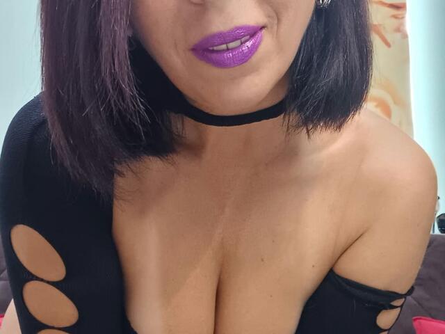 LauraSin with purple lipstick, black choker, and black top with cutouts, showing her large boobs and slightly open top. She has dark brown hair and a confident smile.