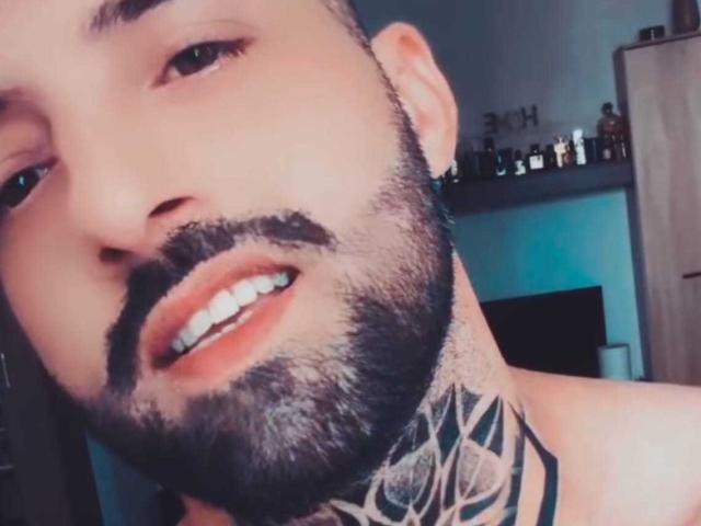 TommyDum with a beard, mustache, and tattoo on the neck, smiling with teeth showing, brown eyes, and ear piercing, in a selfie.