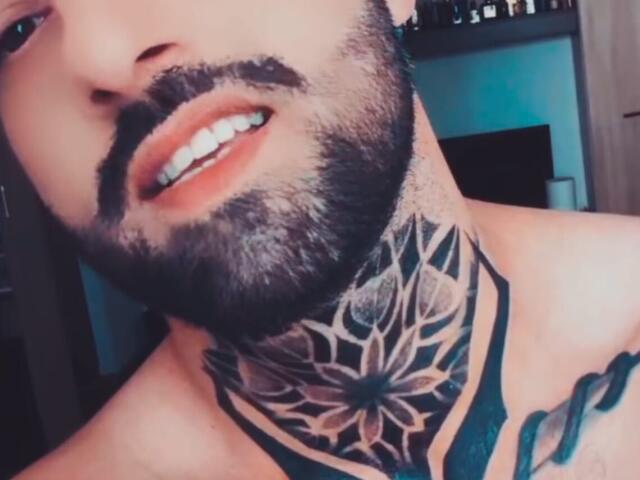 TommyDum ha una barba folta e un tatuaggio a forma di ragnatela sul collo. Le sue labbra sono leggermente socchiuse, mostrando i denti bianchi. Il tatuaggio sul suo petto è parzialmente visibile..