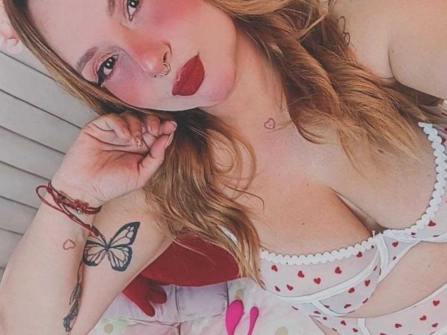 PauleetteRoux a du rouge à lèvres rouge, un piercing au nez et des cheveux blonds ondulés. Elle porte de la lingerie blanche à motifs de cœur, très révélatrice, avec des seins visibles et un tatouage de papillon sur sa main..