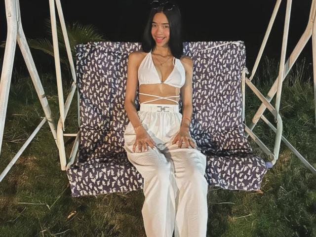 LiaBlom with small tits, wearing a white halter top and high-waisted pants, sitting on a purple and white patterned chair at night, with grass around.