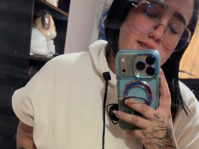 NatalieHarris with glasses, white hoodie, black hair, holding blue phone, tattooed hand, smiling, phone case with ring, camera reflection, medium skin tone, slight nose piercing.