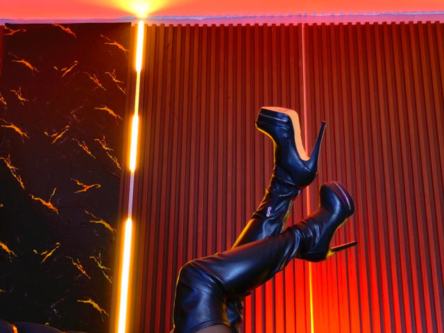 MonroeStone's black leather thigh-high boots with stiletto heels, legs crossed, pointing upward, in a room with red vertical blinds and a glowing yellow line on the left.