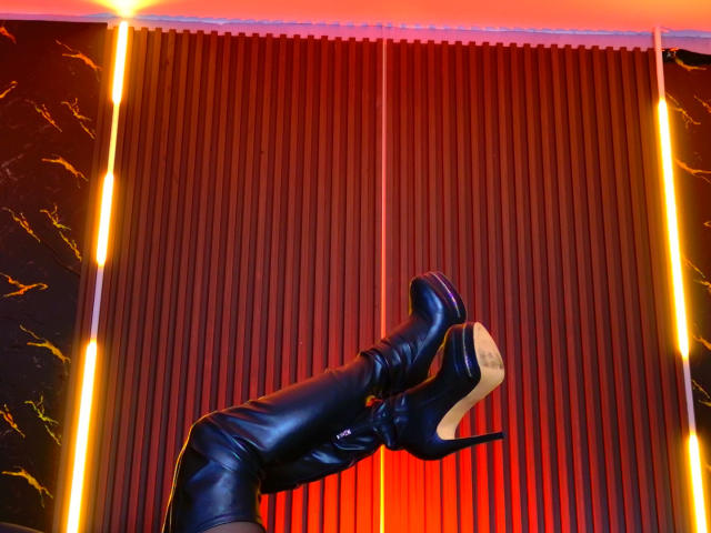 MonroeStone's shiny black thigh-high boots with pointed toes and beige soles are raised in the air, showing the bottom of the heels against a red-lit background.