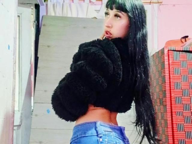 Boorbuja with long black hair, wearing a fuzzy black top and blue jeans, looking over her shoulder, finger in mouth, showing her toned ass.
