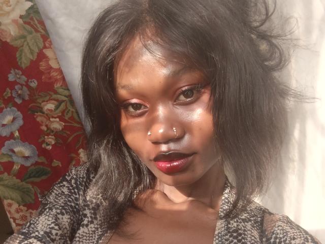 AngelGx has shiny brown skin, dark wavy hair, red lipstick, and a nose piercing. She's wearing a patterned top, looking at the camera with a slight smile.