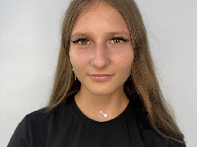 KhadijahSzady tiene ojos verdes, cabello largo y castaño, y lleva una camisa negra con un collar de plata pequeño. Su rostro está ligeramente sonrojado, con una expresión neutra..