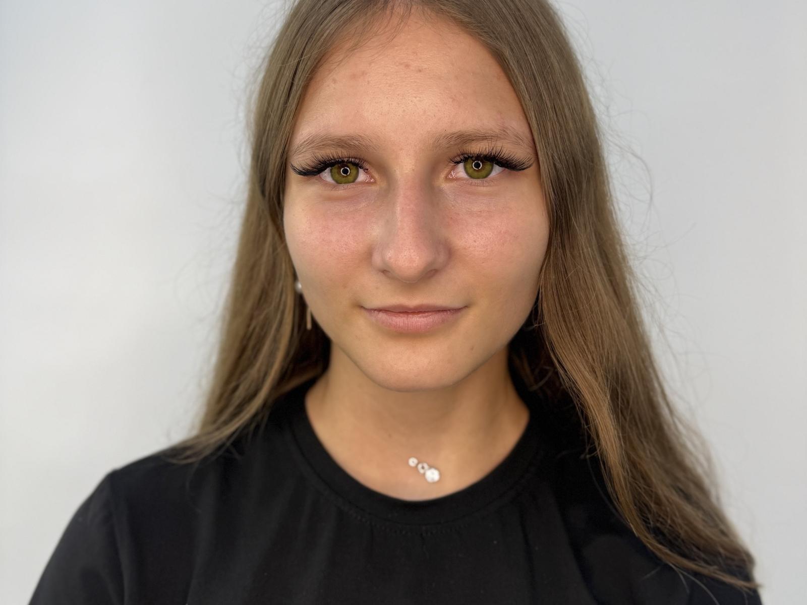 Khadijah Szady hat grüne Augen, langes braunes Haar und trägt ein schwarzes Hemd mit einer kleinen silbernen Halskette. Ihr Gesicht ist leicht gerötet, mit einem neutralen Ausdruck..