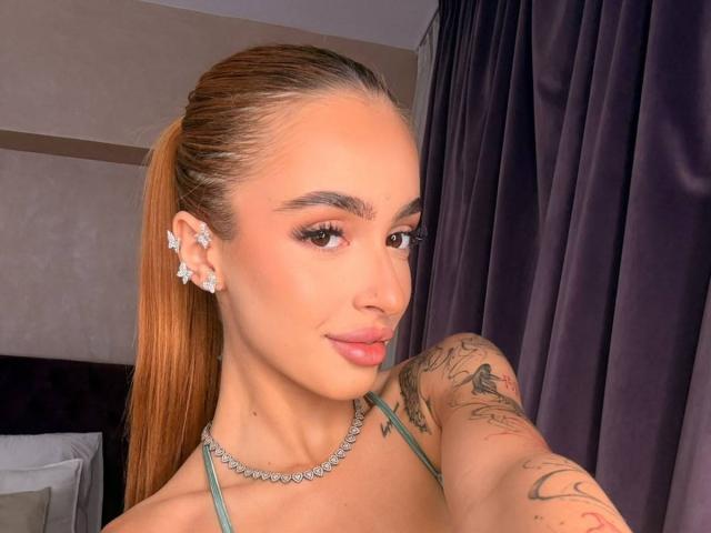 MarissaFex has light skin, brown eyes, and orange ponytail. She's wearing star earrings, a diamond necklace, and a green top, showing tattoos on her right arm.