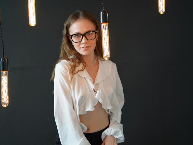 Kristty-hot with glasses, white ruffled blouse, exposed midriff, black panties, standing under hanging Edison bulbs, slight smile, light brown hair, black background.