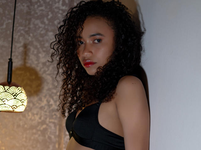ViolettExposito with curly black hair, red lipstick, wearing a black bra, standing against a white wall, light from a patterned lamp illuminating her, looking seductively at the camera.