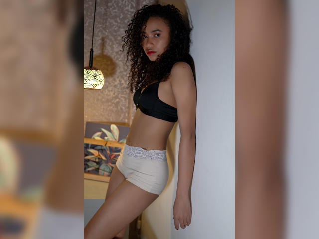 ViolettExposito with curly hair, black bra, and white lace-trimmed panties, standing in a doorway, leaning against a white wall, looking at the camera with a slight pout.