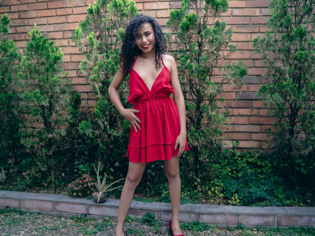 ViolettExposito se tient debout, vêtue d'une robe rouge à décolleté en V plongeant, avec des cheveux noirs bouclés et un sourire. Ses mains sont sur ses hanches, et elle montre sa poitrine et ses fesses, avec un mur de briques en arrière-plan..