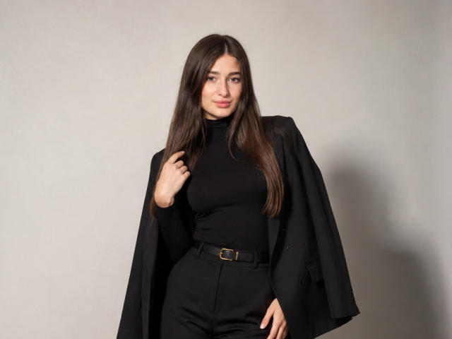 MoonLiza with long brown hair, black turtleneck, black high-waisted pants, and black jacket, holding jacket over shoulder, smiling slightly, hand on hip.