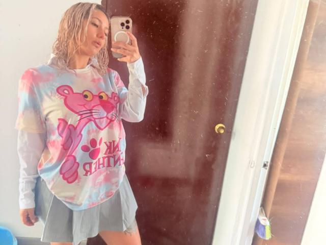 Elena Saenz mit blondierten Haaren, die ein pink-blaues T-Shirt mit einem rosa Bären und einen grauen, gefalteten Rock trägt, macht ein Selfie vor einem Spiegel mit einem rosa Handy..