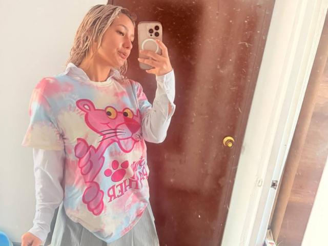 ElenaSaenz with wet blonde hair, wearing a tie-dye pink and blue shirt with a cartoon cat, holding a phone, in front of a mirror. She's smiling.