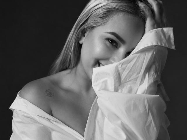 Black-and-white photo of AshleyRossie with long blonde hair, smiling, wearing an off-shoulder white shirt, exposing a small tattoo on her right shoulder. Her hand's in her hair.