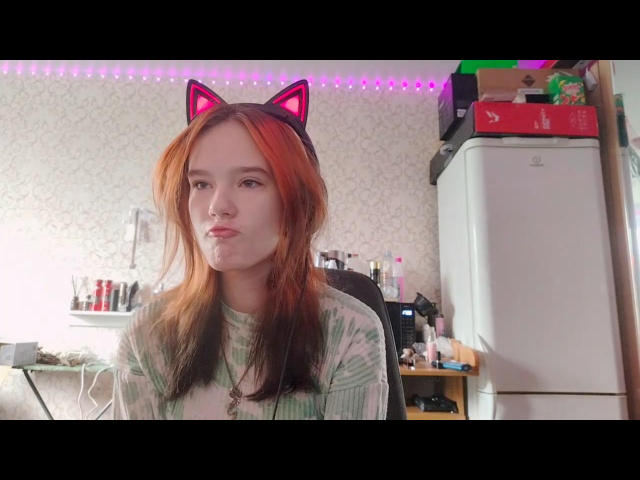 RosalieBayers with red and brown hair, wearing cat ear headband, pouty face, green striped shirt, in a room with a fridge and shelves, looking slightly bored.