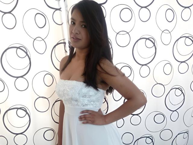ArianithaXox stands in a white dress with lace detail, her dark hair flowing, hand on hip, looking at the camera with a slight smile, showing her tits.