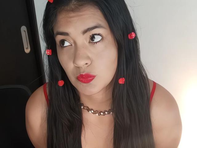 ArianithaXox with long black hair, red lipstick, and red clips in her hair, wearing a red top and a brown necklace, pouting her lips. Brown eyes, looking sideways.