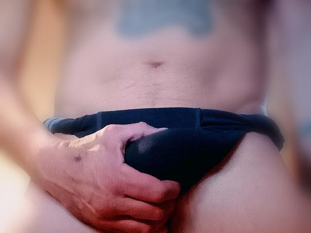 La mano di Malino69 stringe della biancheria intima nera sopra il suo pene flaccido e circonciso. Si vede l'ombelico, un tatuaggio sopra la vita, immagine leggermente sfocata. La pelle di Malino69 è chiara, la mano è muscolosa..