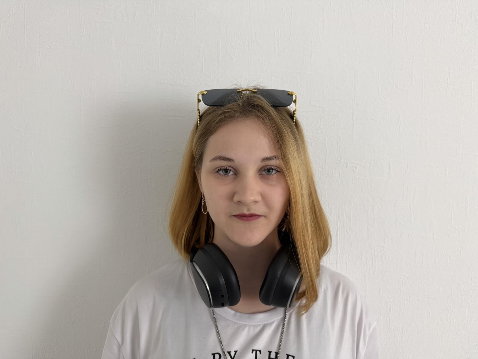 Carissa Cariddi, con i capelli biondi, indossa una camicia bianca, ha delle cuffie nere intorno al collo e degli occhiali da sole in testa, ha un'espressione neutra e si trova in una fotografia semplice..