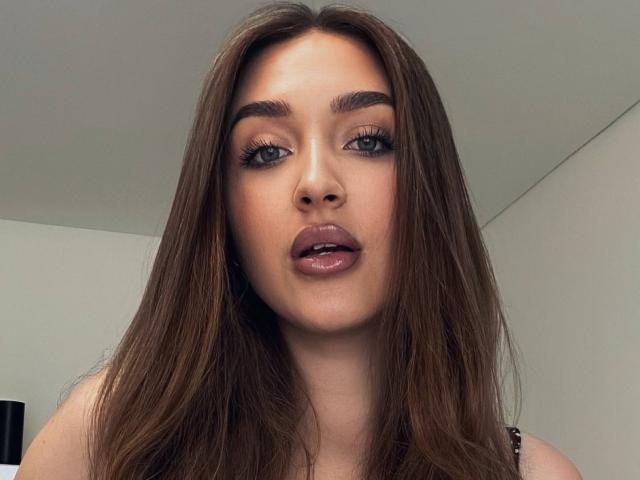 HeePickman has long brown hair, thick eyebrows, green eyes, and glossy pink lips. Her makeup includes long lashes and subtle eyeshadow. She's looking at the camera with a slightly open mouth.