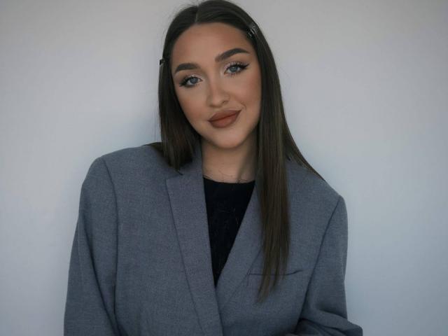 HeePickman with long brown hair, grey blazer, black top, and subtle makeup, smiling softly. HeePickman's tits are not visible. HeePickman's ass is not visible. HeePickman looks cute.